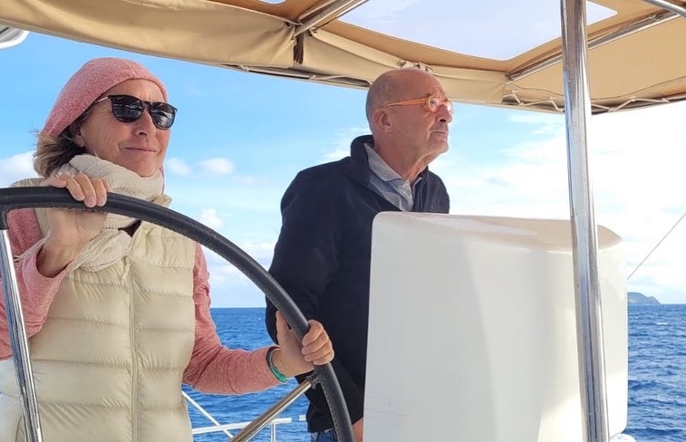 At the helm with your partner. This sailing woman is smiling. She wears sunglasses while her male partner looks somewhere else.