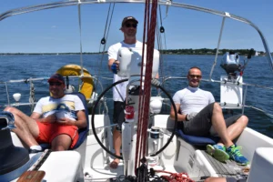 thre men on a sailboat. one man is at the helm while the two others are sitting and smiling