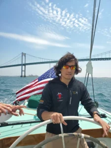 A blind veteran at the helm of an America's cup boat with Newport bridge in the background