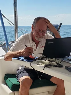 Tom at a table on board his catamaran. scratching his head in front of his laptop before living on board